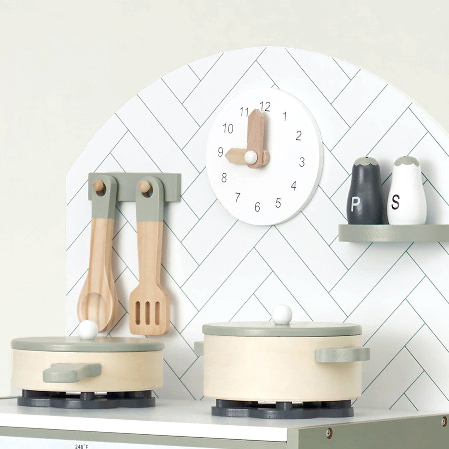 Children's kitchen playset with pots, pans, and utensils on a white surface against a white wall with a clock.