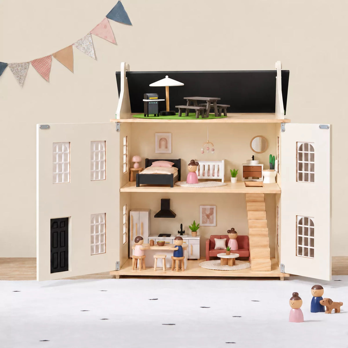 A dollhouse-style house with open doors and windows, shown fully furnished with miniature furniture, bedding, kitchen, and outdoor seating. The dollhouse is photographed in a styled room setting with a beige wall, bunting banner, and a few small toy figures on the floor.